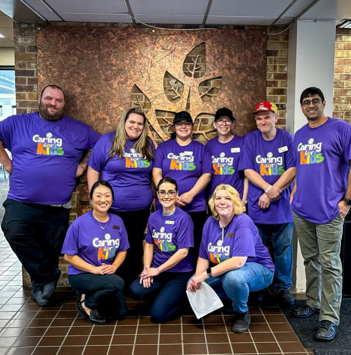 a group of people in purple Caring for Kids shirts standing next to a brick wall