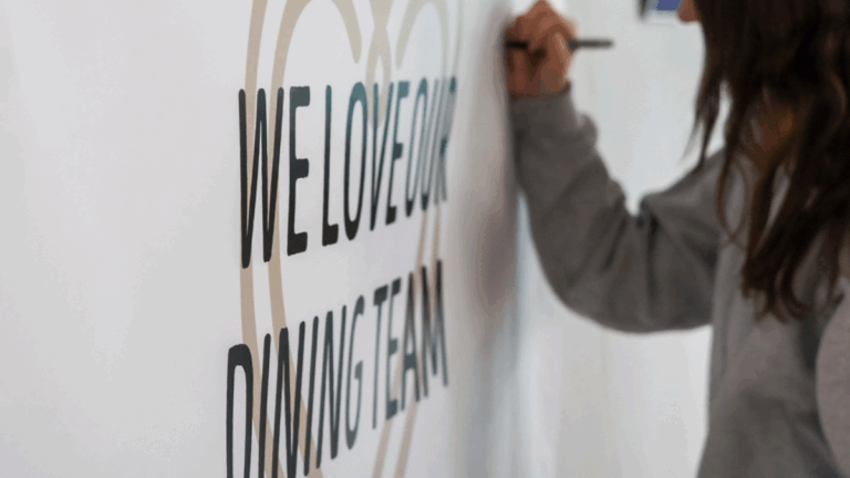 a female college student writes a message on a sign that says We Love Our Dining Team