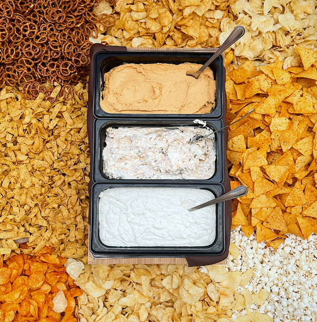 three dips surrounded by pretzels, chips, and more snacks